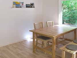 A wooden dining table with four chairs, a window with a view of greenery, and a shelf with various items.