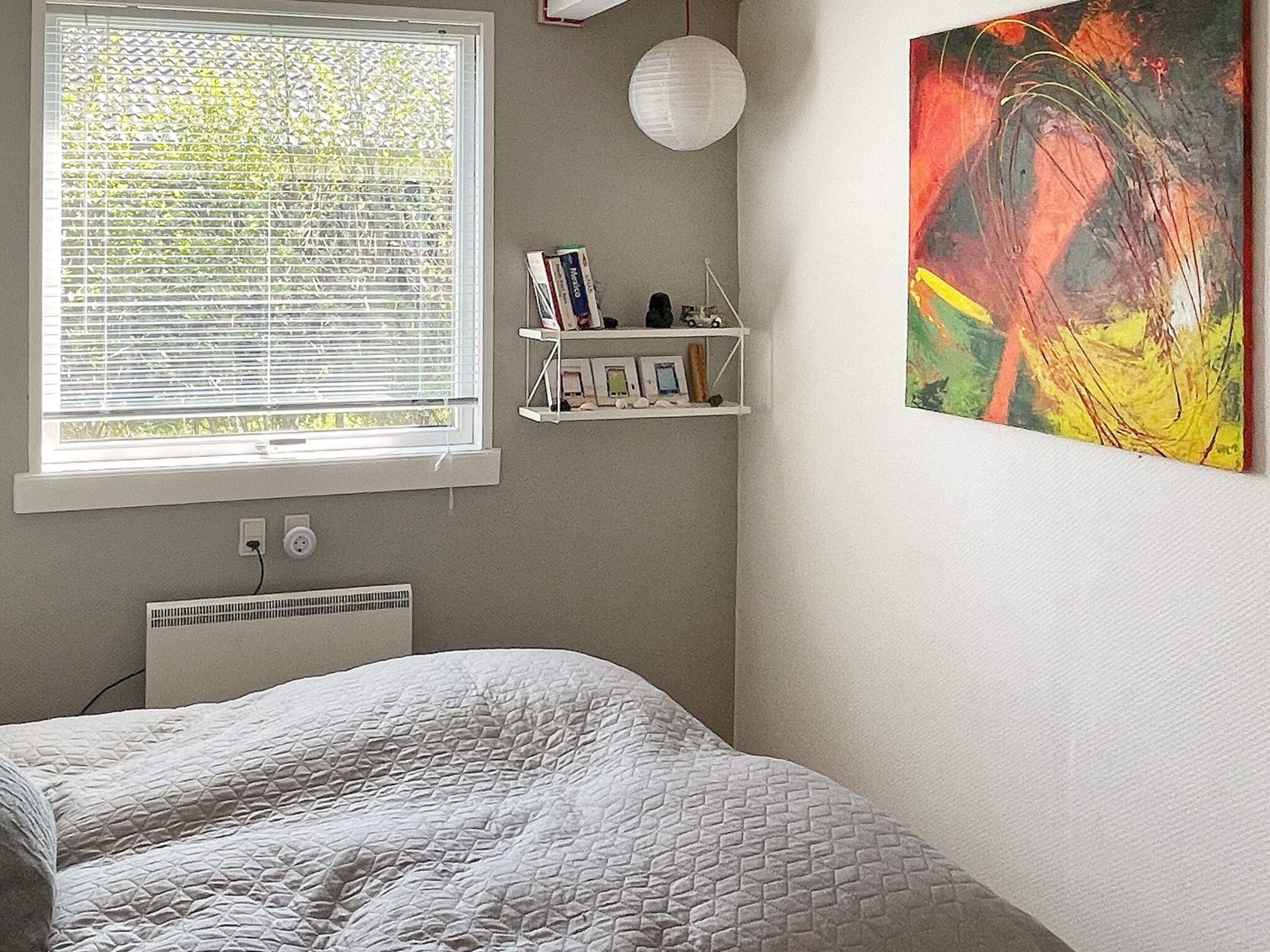 Ein Schlafzimmer mit einem Bett, einem Fenster mit Jalousien, einem Heizkörper, einem Regal mit Büchern und Bildern sowie einem Gemälde an der Wand.