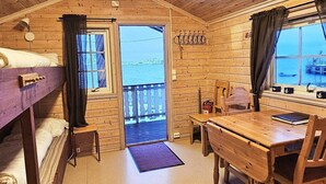 En stuga med en våningssäng, ett skrivbord i trä, en stol, ett litet bord, ett fönster med gardiner och utsikt över vattnet.