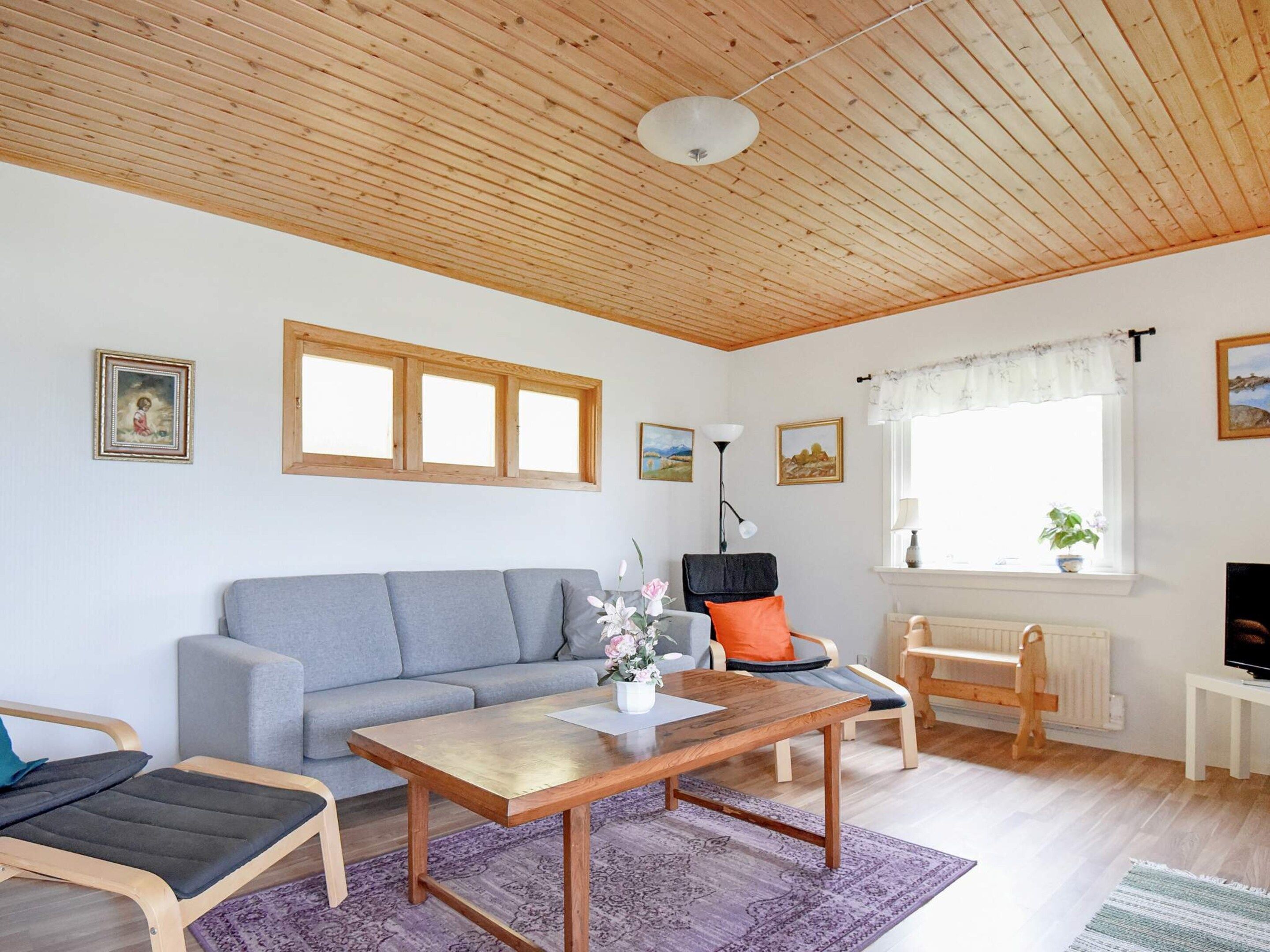 A living room with a wooden ceiling, a grey sofa, a wooden coffee table, a chair, a small table with a lamp, and a window with a plant.