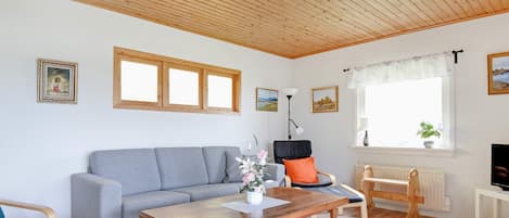 A living room with a wooden ceiling, a grey sofa, a wooden coffee table, a chair, a small table with a lamp, and a window with a plant.