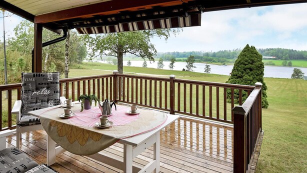 Une terrasse extérieure couverte, équipée d’une table pour deux, entourée de rampes en bois et offrant une vue sur un lac.