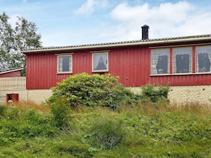 A red wooden house with three windows, a chimney, and a small bush in front.
