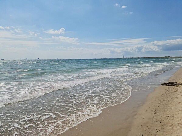 Una playa con olas, cielo despejado y barcos a lo lejos.