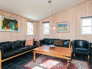 A room with a black leather sofa, a wooden coffee table, and a black leather armchair. There are two windows with wooden frames and a painting on the wall. - 4 Star Holiday Home in Ebeltoft (Ebeltoft)