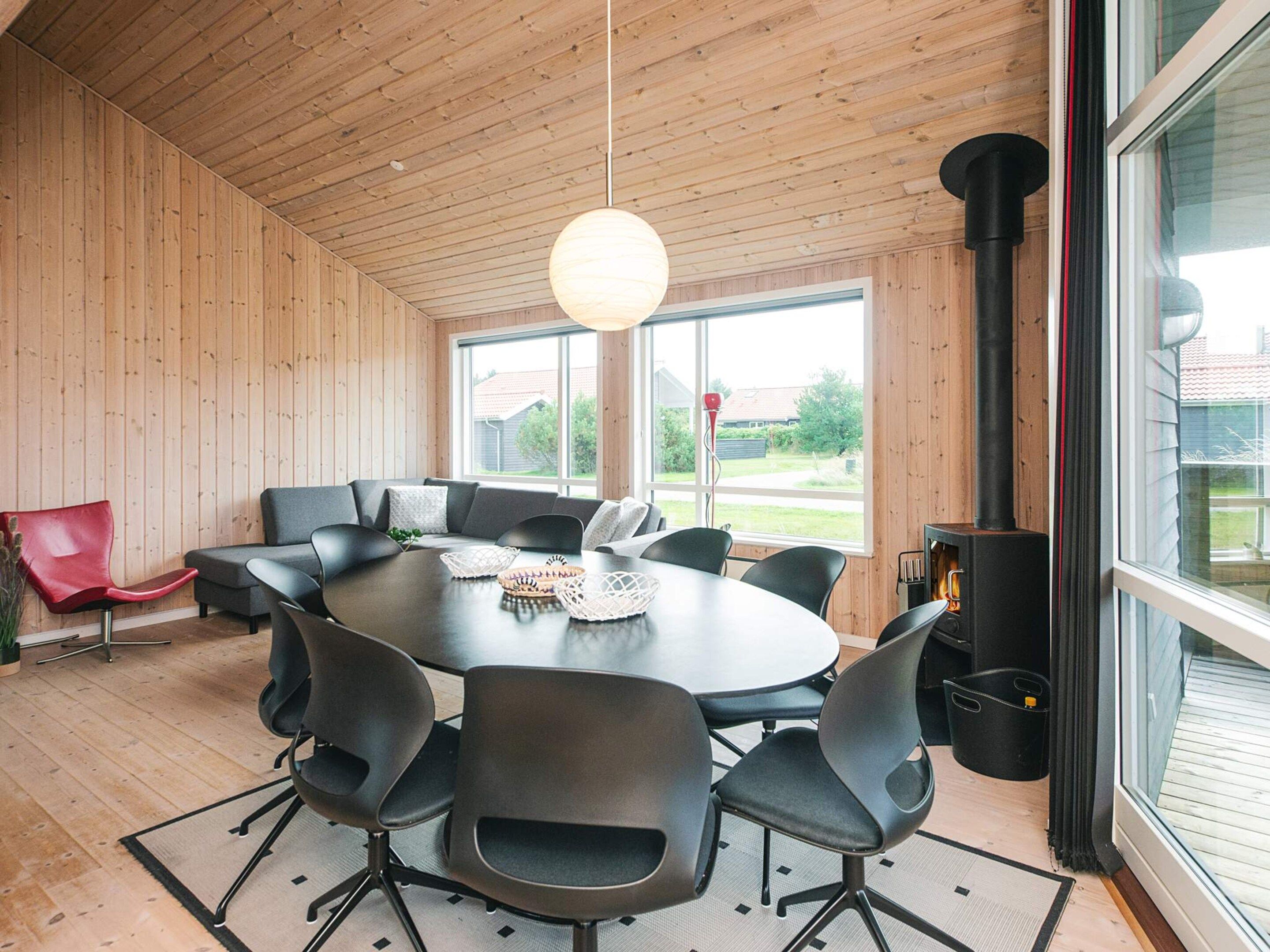 A modern interior with a round table surrounded by chairs, a red chair, a sofa, and a large window with a view of the outside.