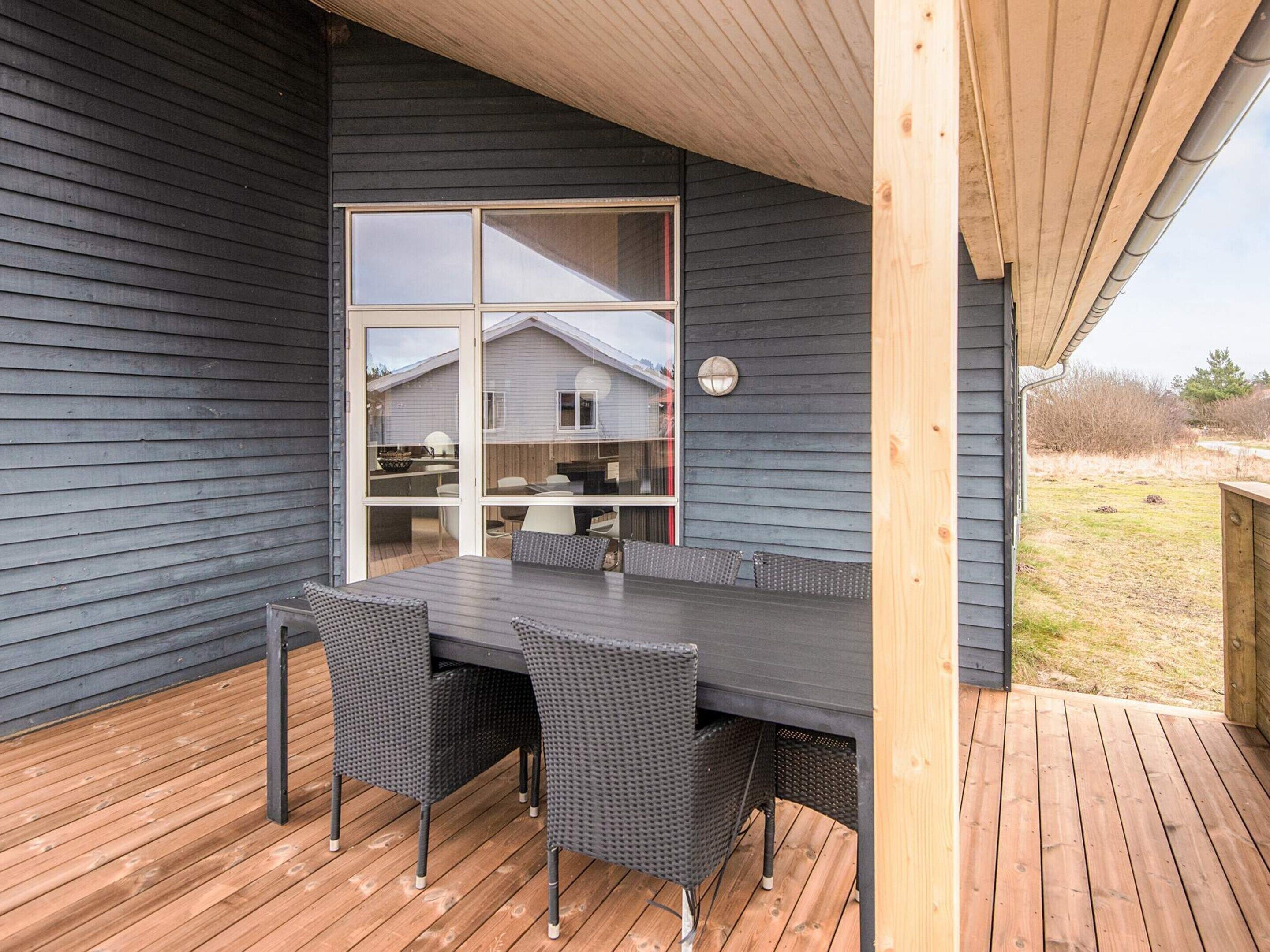 Un espacio exterior techado con mesa y sillas, vista a una casa a través de una puerta de vidrio y una terraza de madera.
