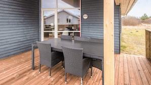 A covered outdoor dining area with a table and chairs, a view of a house through a glass door, and a wooden deck. - Cozy Holiday Home in Ulfborg near Sea (Ulfborg)