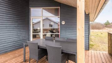 Un espacio exterior techado con mesa y sillas, vista a una casa a través de una puerta de vidrio y una terraza de madera.