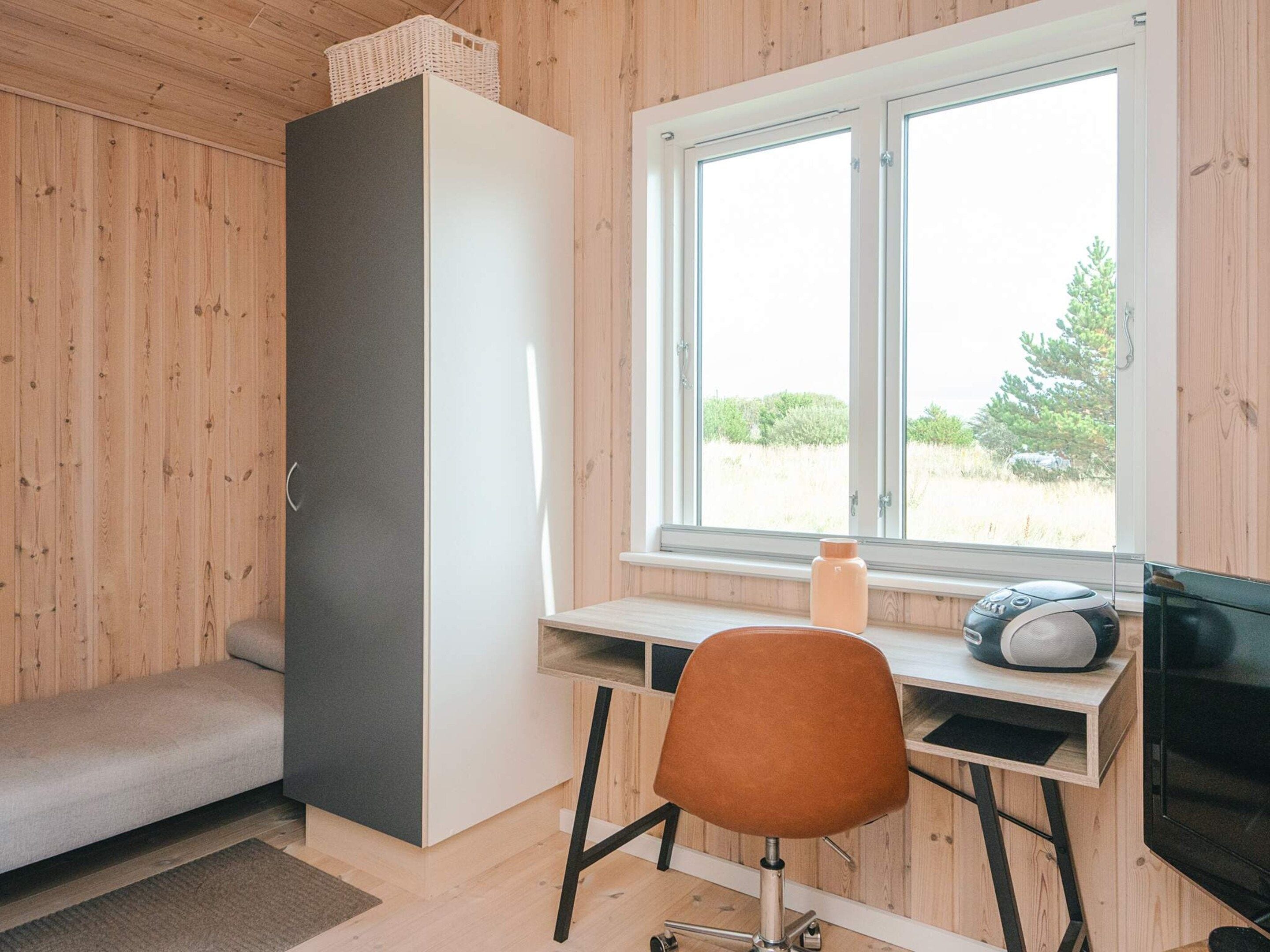 Ein Zimmer mit einer Holzwand, einem weißen Schrank, einem Schreibtisch mit Stuhl und einem Fenster mit Blick auf Grünflächen.