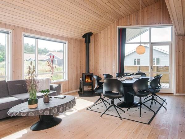 A modern interior with a wooden ceiling, a fireplace, a dining area with chairs, and large windows.