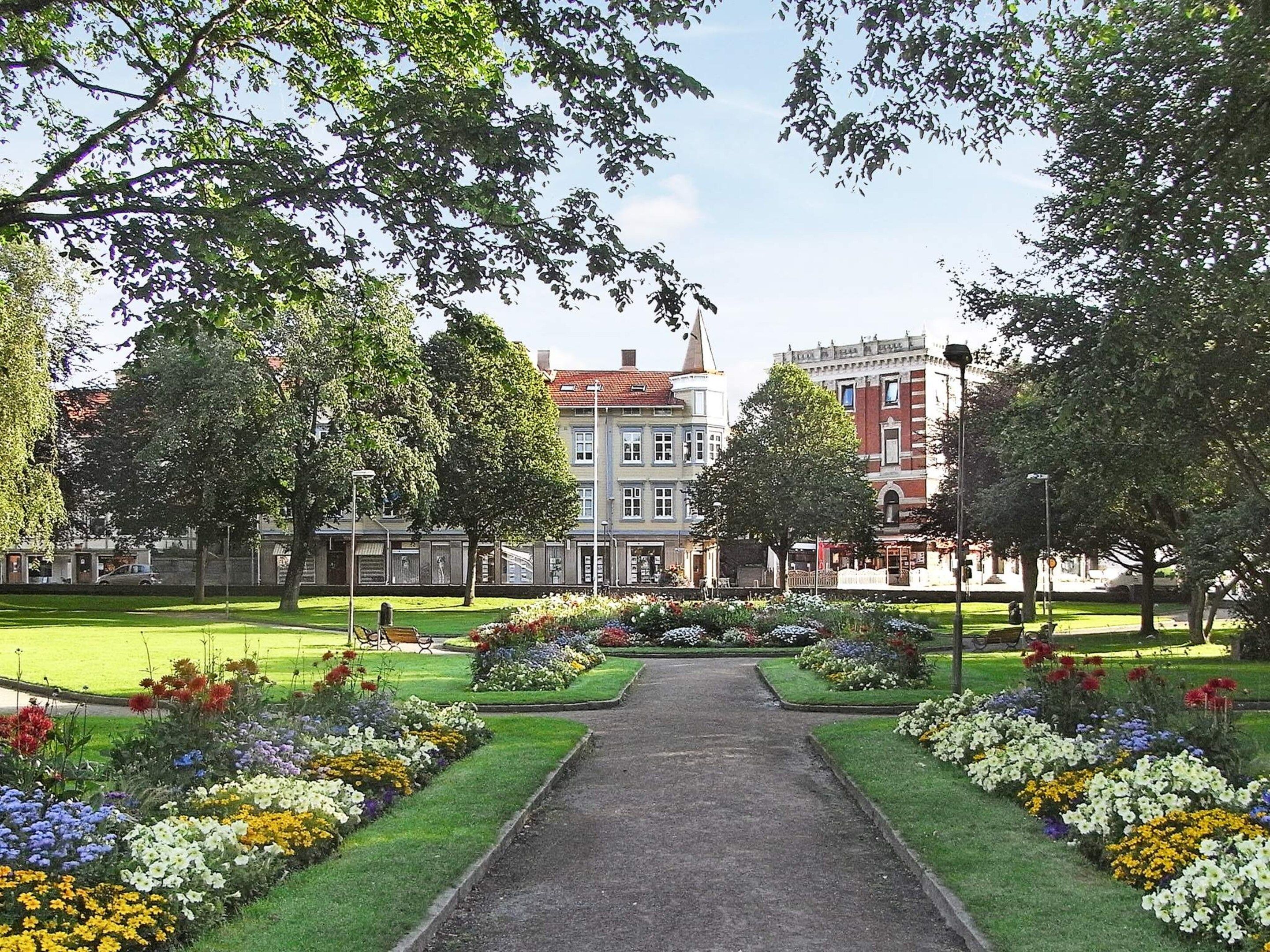 Um caminho serpenteia por um parque, com canteiros de flores de ambos os lados, cercado por árvores e edifícios ao fundo.