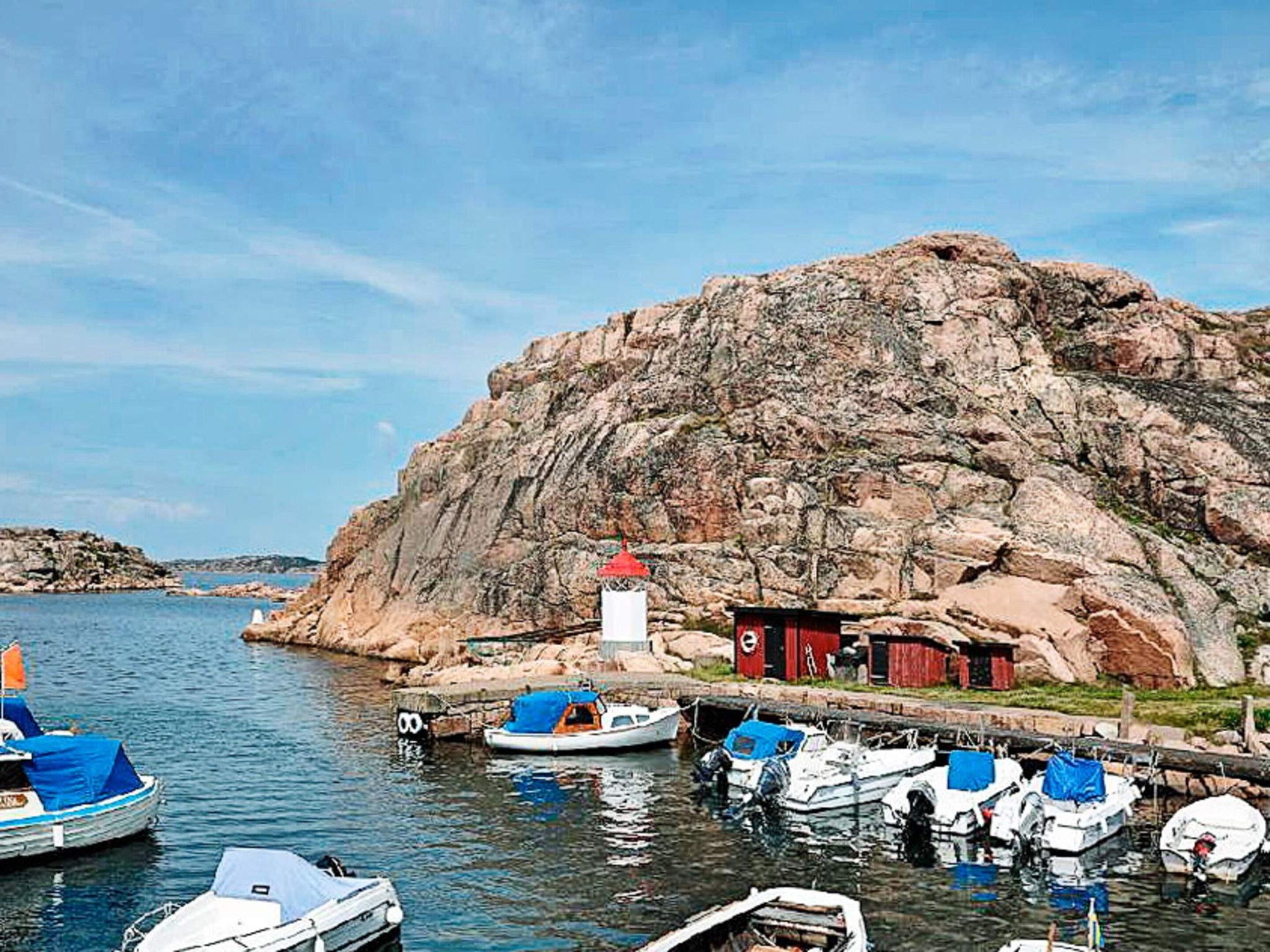 Um litoral rochoso com barcos ancorados em um cais, um farol e um pequeno edifício vermelho.