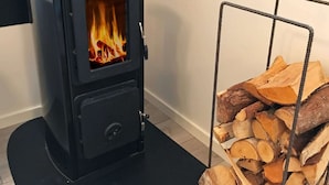 A black wood-burning stove with visible flames, a metal rack with cut logs, and a tiled floor. - 4 Star Holiday Home in Akersberga (Akersberga)