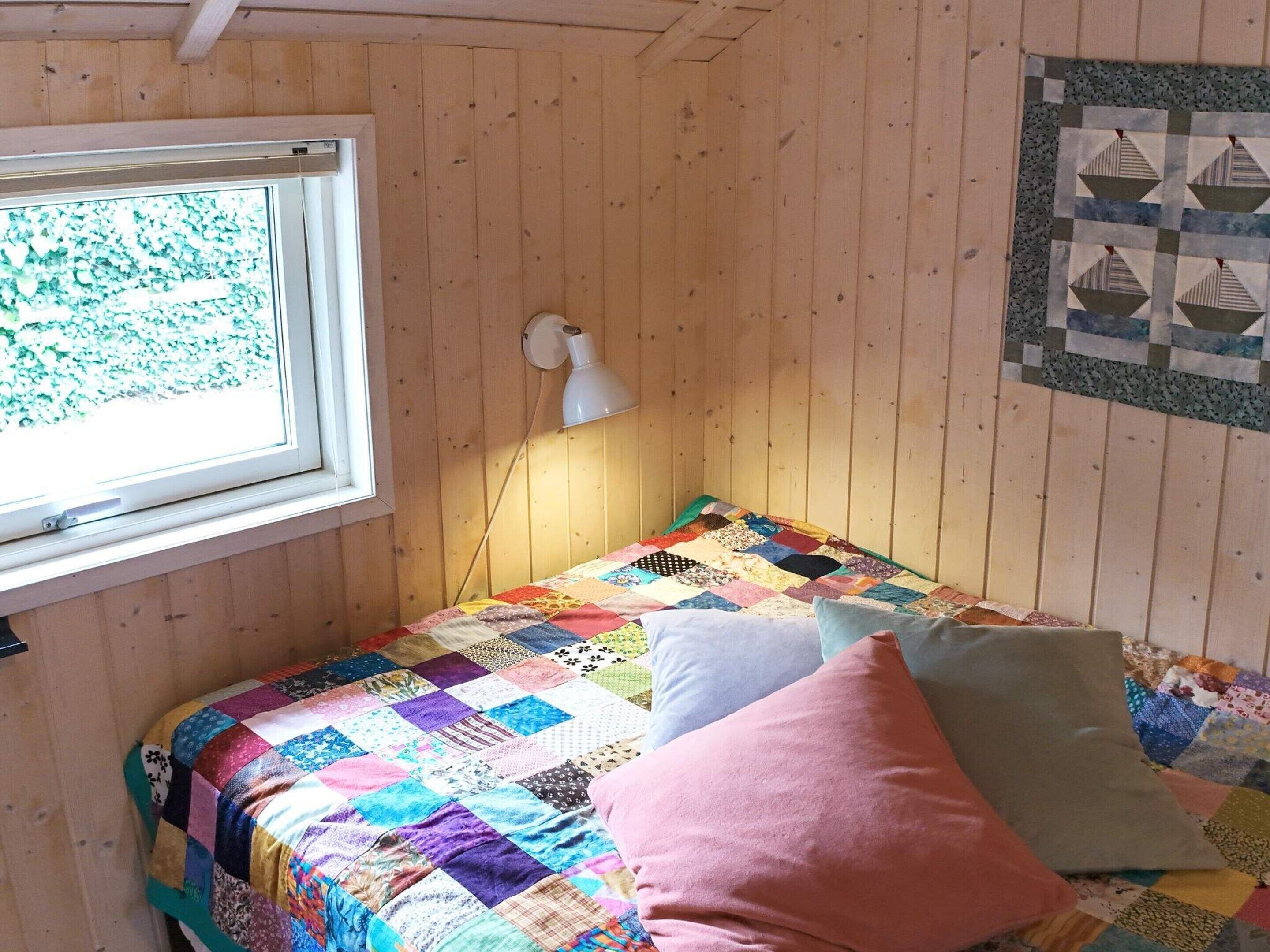 A cozy room with a wooden interior, a bed with a colorful quilt, a white lamp, and a window with a view of trees.