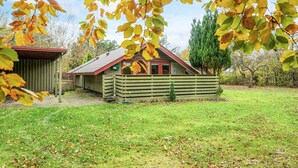 Une cabane en bois avec une porte rouge et un toit en pente, entourée d’une cour verdoyante et d’arbres.