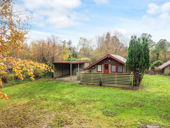 A rustic cabin with a wooden fence, a covered area, and a small tree in a grassy yard.