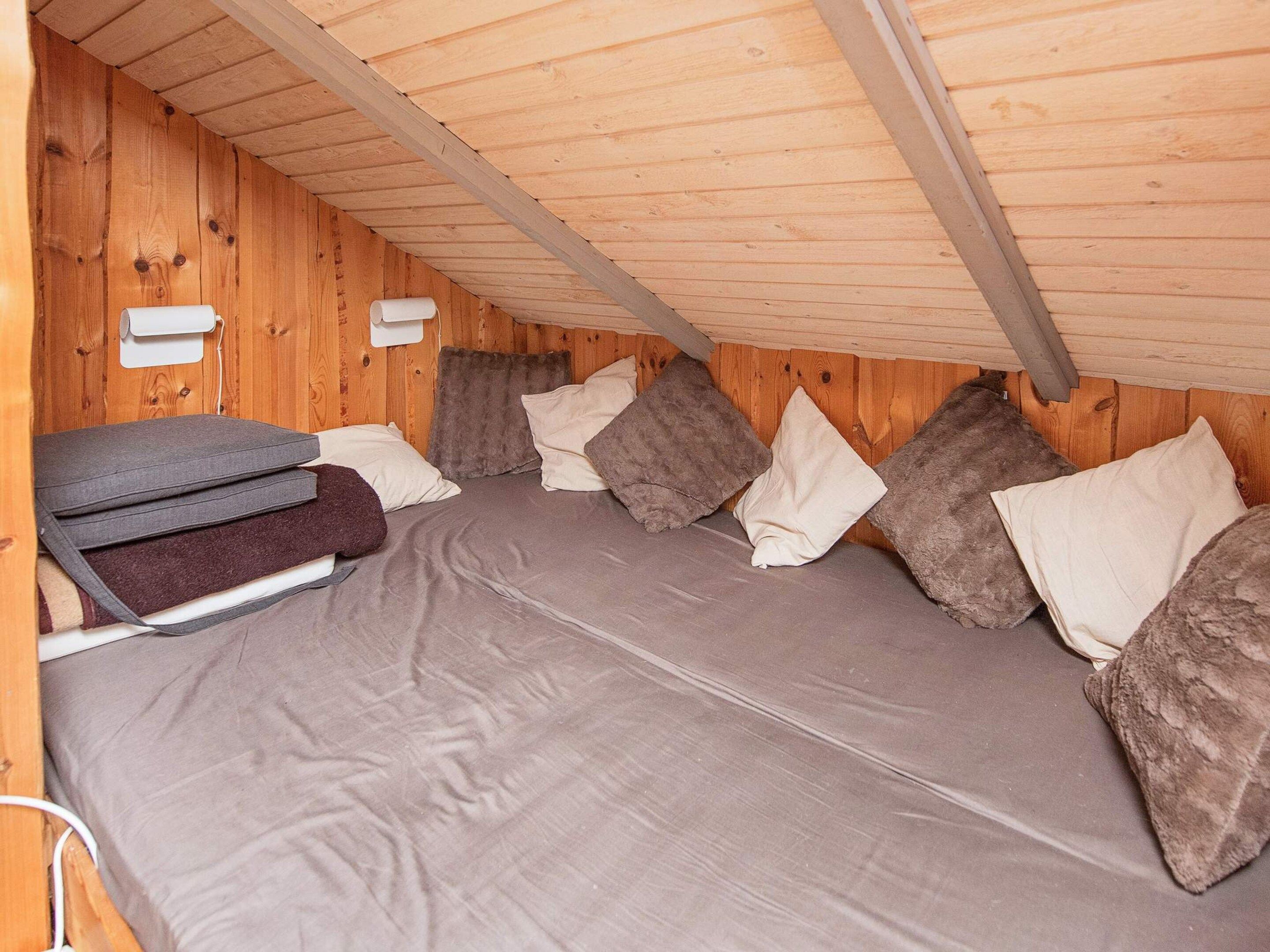 A compact room with a sloped ceiling, wooden walls, and a bed with grey bedding and multiple pillows.