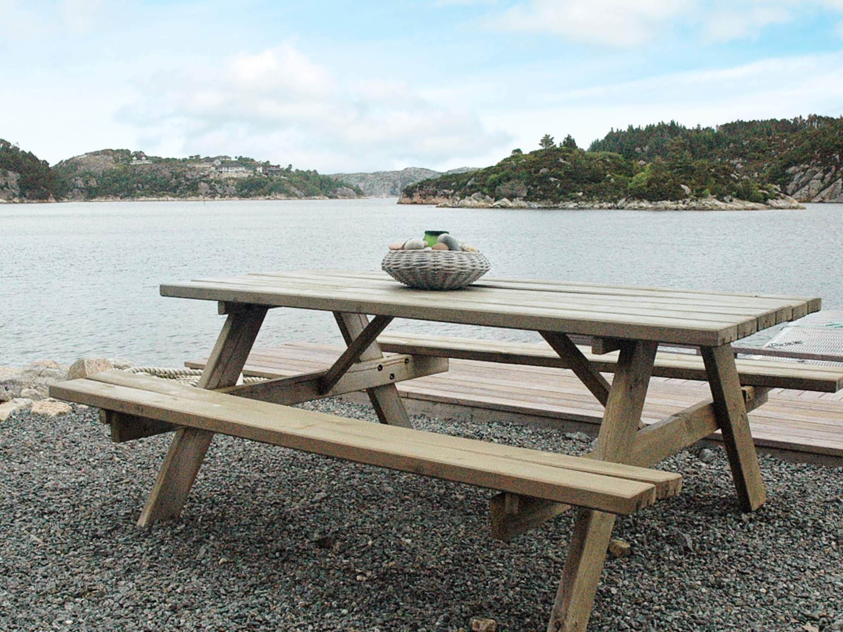 A wooden picnic table with benches on a pebbled shore by a lake, with hills in the background.