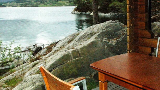 Een houten terras met tafel en stoelen met uitzicht op een rustig meer, omgeven door rotsachtig terrein en begroeiing.