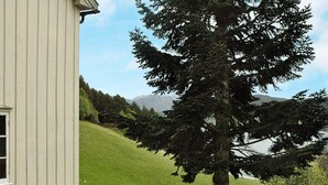 A white building with a porch, a large evergreen tree, and a view of a grassy hill with mountains in the distance. - 5 Person Holiday Home in Vagland (Heim)