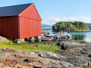 A red wooden building with a sloped roof, situated by a rocky shoreline with boats docked nearby. - 5 Person Holiday Home in Vagland (Heim)