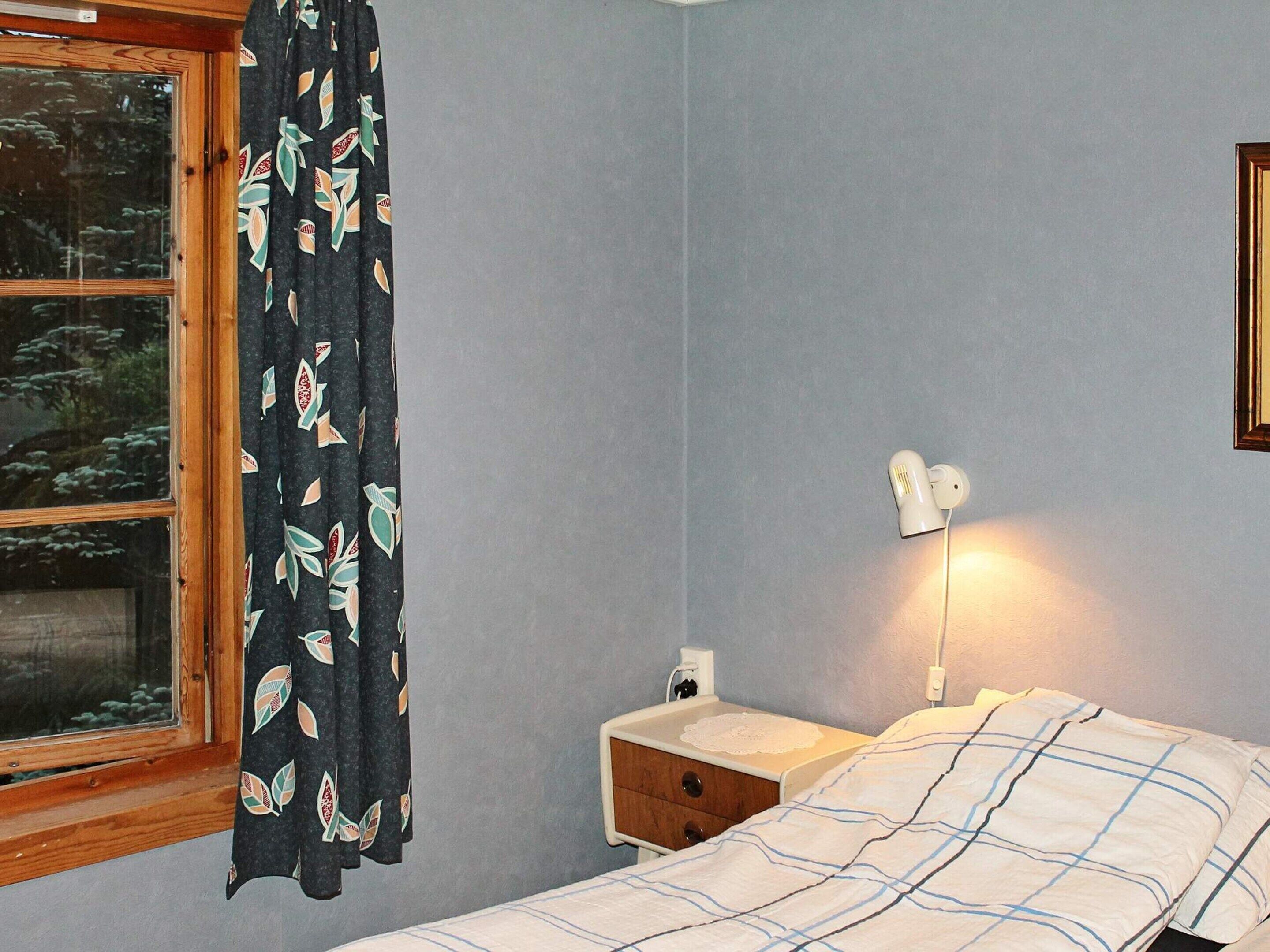 A bedroom with a bed, a nightstand, a lamp, a window with curtains, and a wall-mounted light.