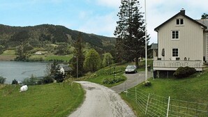 A rural road leading to a two-story house with a porch, surrounded by greenery and a lake. - 5 Person Holiday Home in Vagland (Heim)