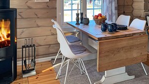 Interior acogedor de una cabaña con una mesa de comedor de madera, sillas blancas, una chimenea y un ventanal con vistas al exterior.