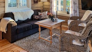 Un acogedor interior de cabaña de madera con un sofá de color oscuro, un sillón de mimbre, una mesa de madera con un jarrón de flores y un televisor en la pared.