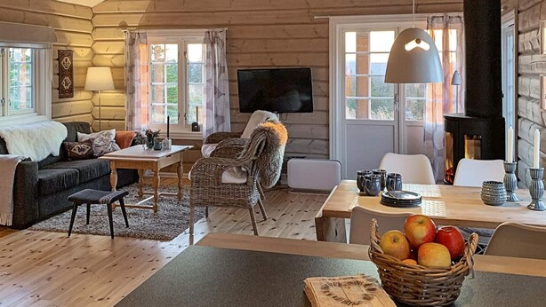 Interior acogedor de una cabaña con pared de troncos de madera, chimenea, televisor, mesa de comedor con manzanas y zona de estar cómoda.