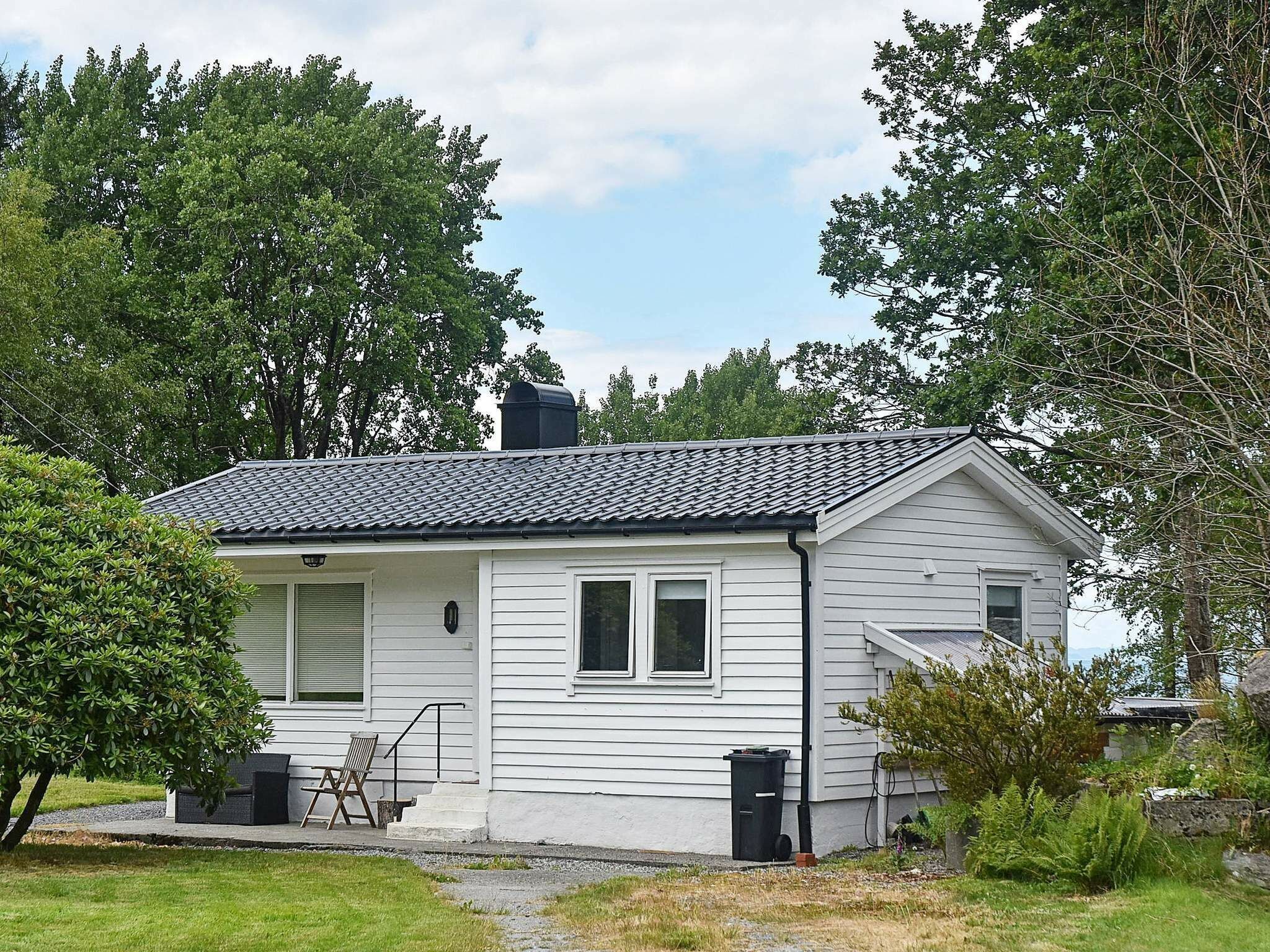 A single-story house with a tiled roof, white siding, and a chimney. There is a small porch with a chair and a trash can.