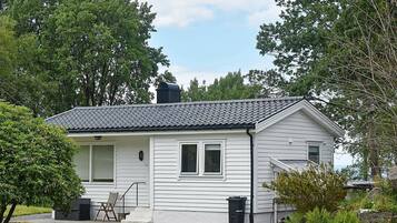 A single-story house with a tiled roof, white siding, and a chimney. There is a small porch with a chair and a trash can.