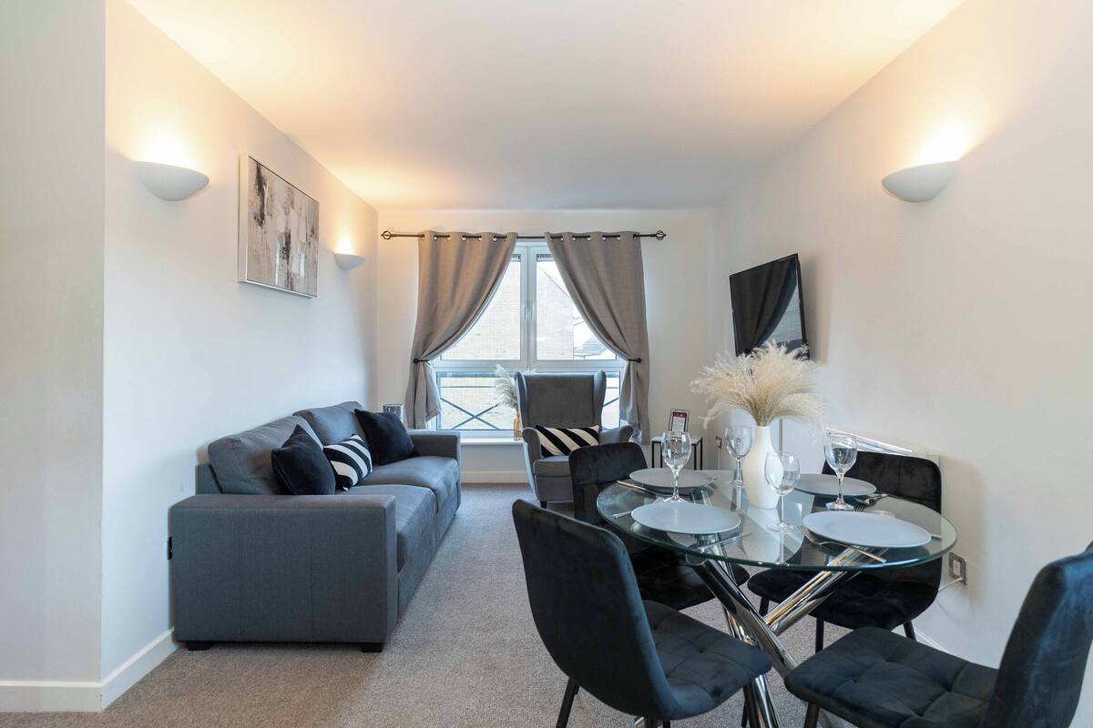 Un salon moderne avec un canapé gris, des coussins à rayures noires et blanches, une table à manger en verre avec des chaises, et une fenêtre avec des rideaux.