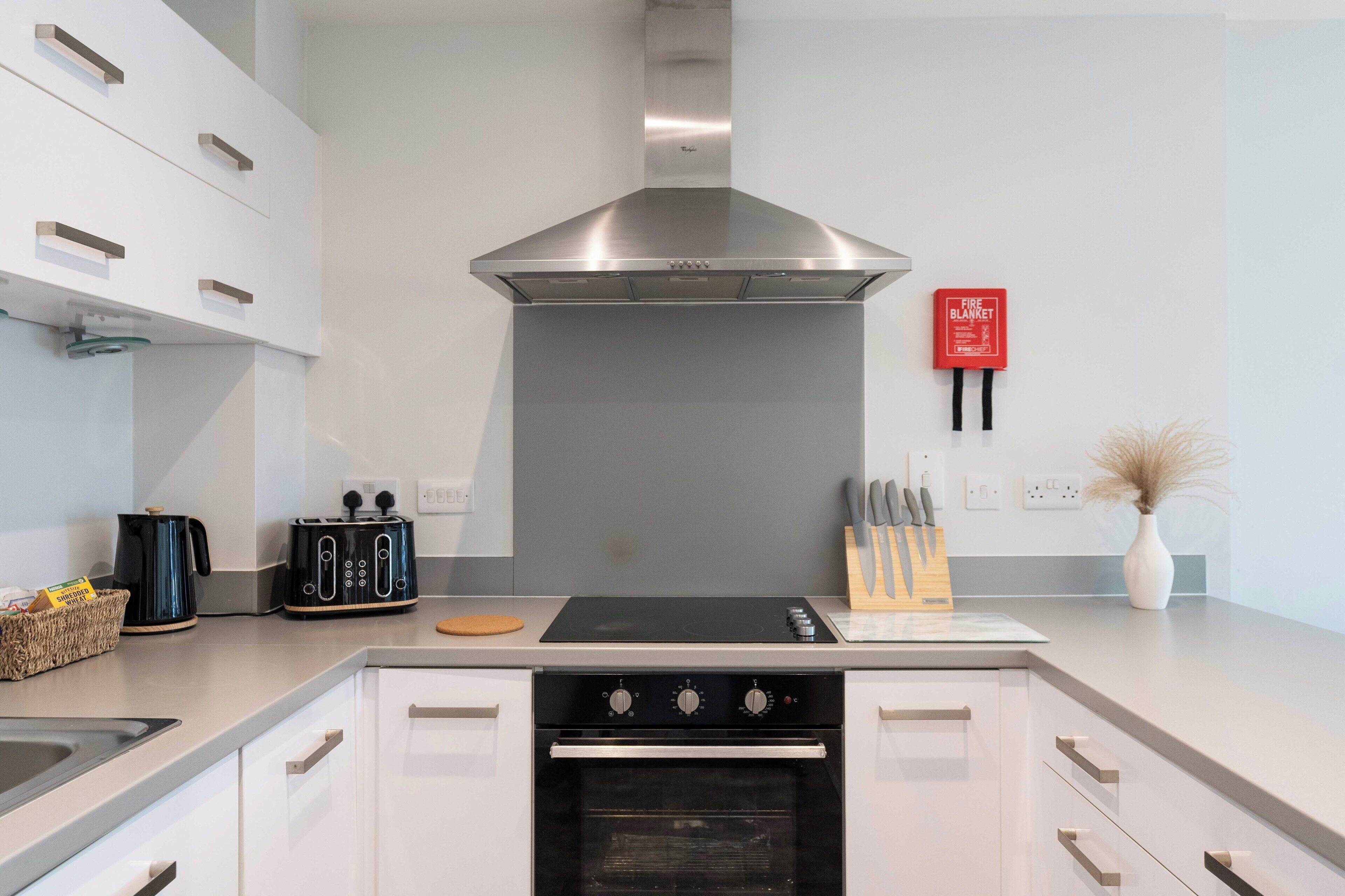 A modern kitchen with white cabinets, a stainless steel range hood, a built-in oven, and a fire extinguisher on the wall.