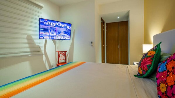 A hotel room with a bed, a television mounted on the wall, a wooden door, and colorful pillows.