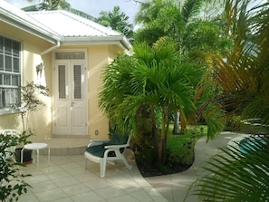 A small, yellow building with a white door and windows, surrounded by lush greenery and a tiled patio area with a table and chairs.