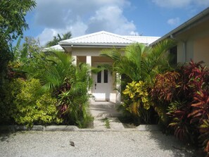 A house with a white roof and columns, surrounded by tropical plants and a gravel pathway leading to the entrance. - Cherry Garden Villa Apartment - private pool in desirable Rockley Beach location (Rockley, Christ Church)