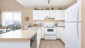 A modern kitchen with white cabinets, a stove, a microwave, and a refrigerator. There is a window with blinds and a plant on the counter.