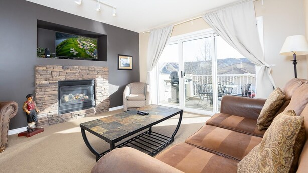 A living room with a stone fireplace, a television mounted above it, a sofa, a coffee table, and a view through a glass door to an outdoor patio. - Paradise Canyon Golf Resort - Luxury Condo U409 (Lethbridge)