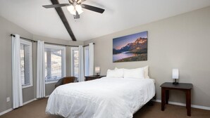 A hotel room with a large bed, a ceiling fan, a painting of a landscape, and a view of the outdoors through the windows.