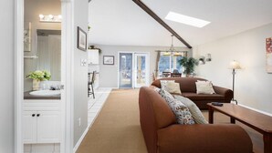 A spacious living area with a brown sofa, a wooden coffee table, and a lamp. There is a white cabinet with a sink and a mirror. A door leads to a balcony with a view of the outdoors.