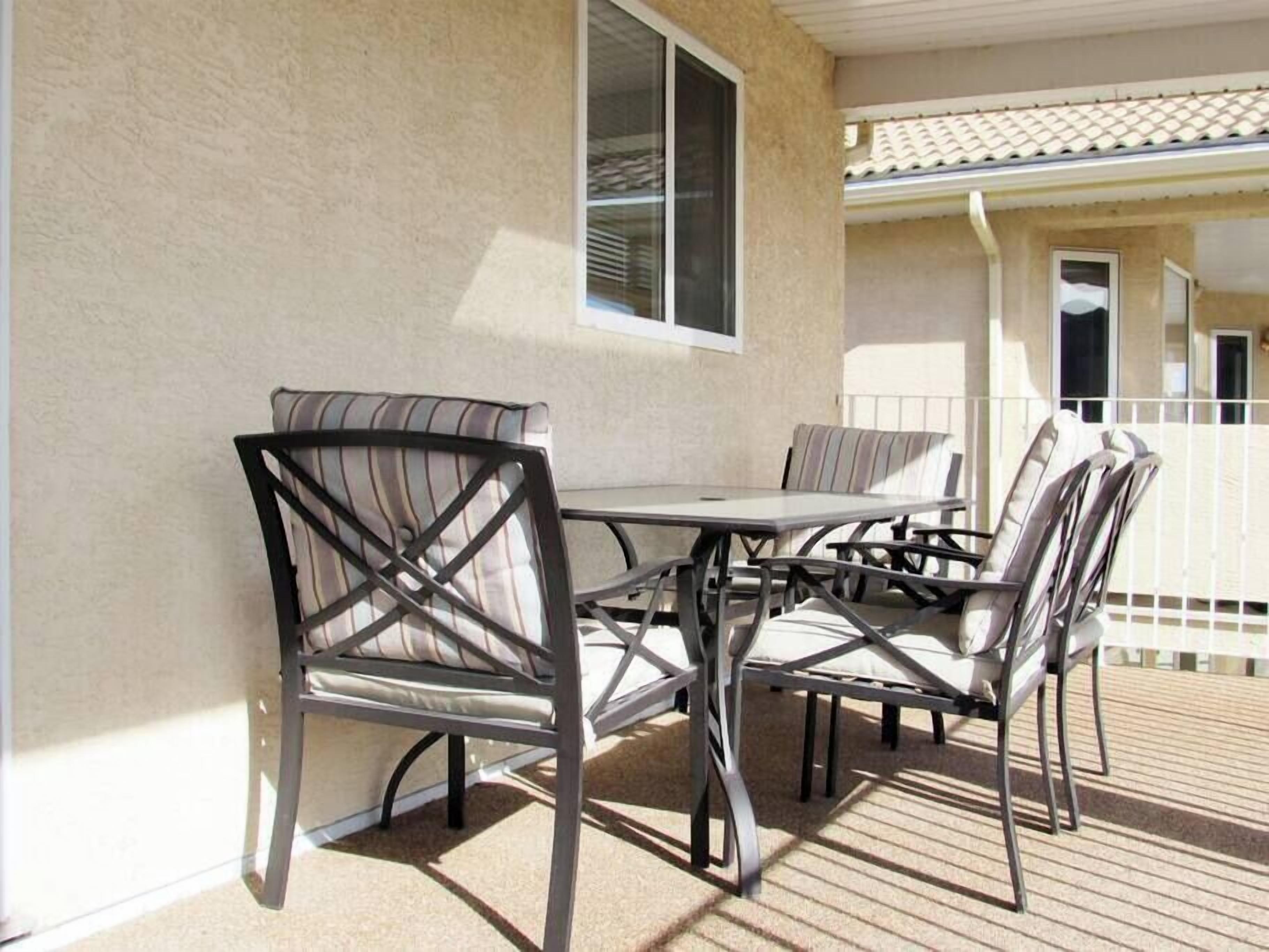 Um pátio externo com mesa e cadeiras, corrimão branco e um edifício com telhado de telhas ao fundo.