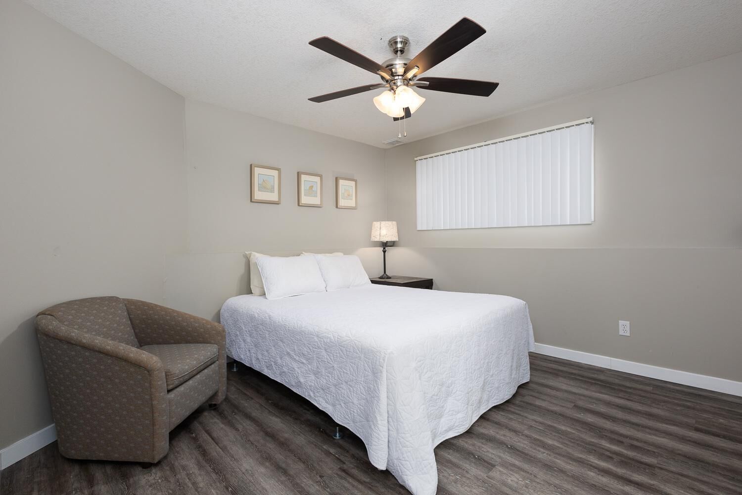 Une chambre à coucher comprenant un lit, une chaise, un ventilateur de plafond, une table de chevet et une fenêtre avec des stores.
