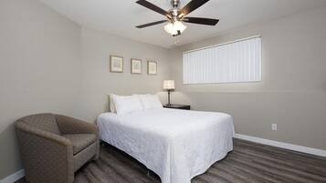 Une chambre à coucher comprenant un lit, une chaise, un ventilateur de plafond, une table de chevet et une fenêtre avec des stores.