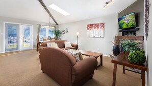 A spacious living room with a brown sofa, a wooden coffee table, and a flat-screen TV mounted on the wall. There is a dining area with a table and chairs, and a large window offering a view of the outdoors. - Paradise Canyon Golf Resort - Signature Villa 380 (Lethbridge)