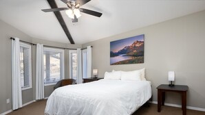A hotel room with a large bed, a ceiling fan, a painting of a mountain landscape, and a view of the city through the windows. - Paradise Canyon Golf Resort - Signature Villa 380 (Lethbridge)