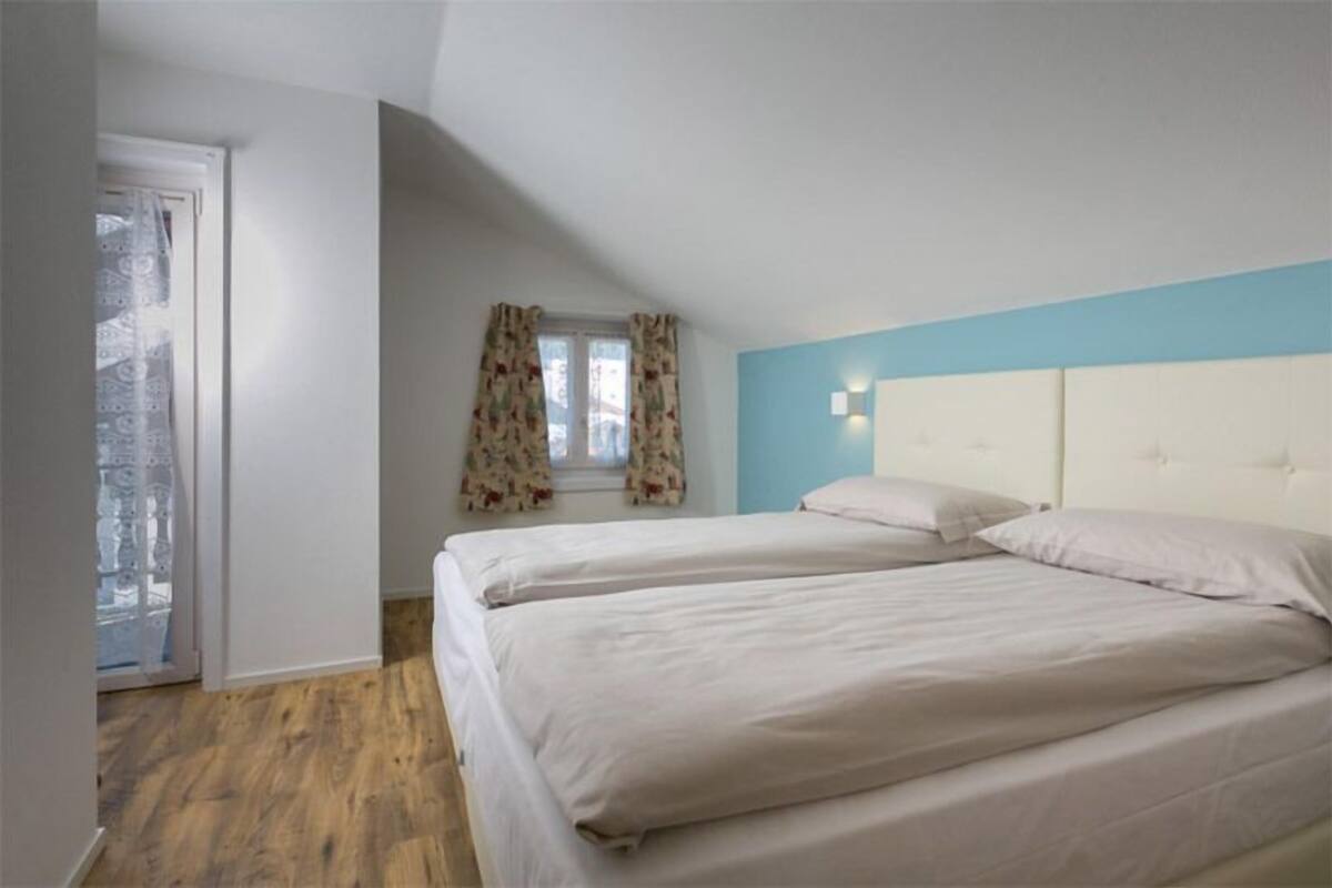 A room with two beds, a window with floral curtains, and a light blue wall.