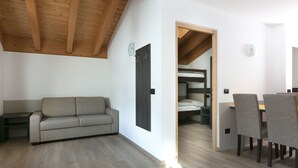 A modern room with a wooden ceiling, a sofa, a dining area with chairs, and a bunk bed in the background.