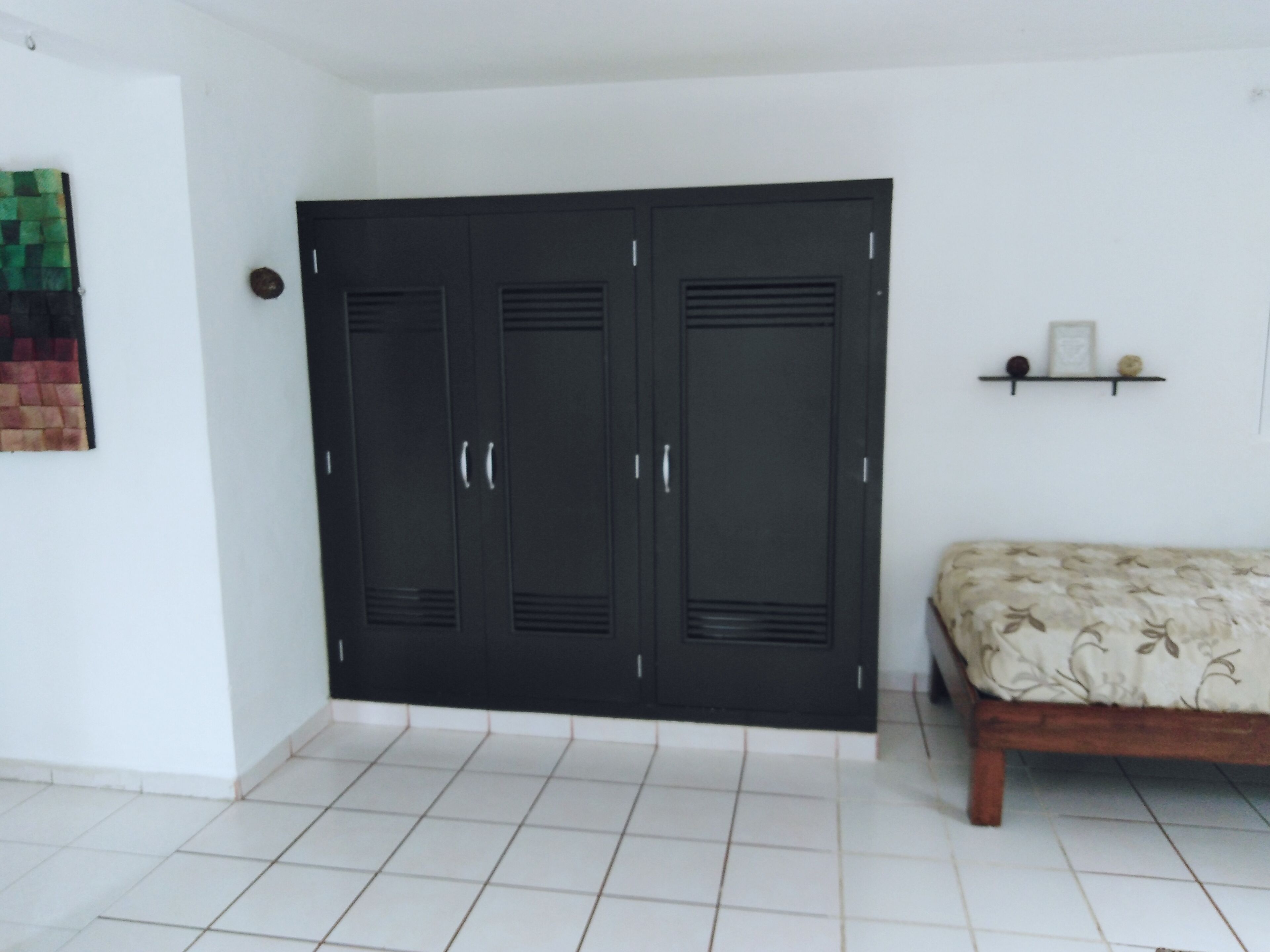 A room with a large black wardrobe, a bed with a patterned bedspread, and a small shelf with decorative items.